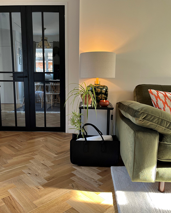 Corner of stylish living room with green fabric sofa, golden coloured herringbone flooring and black blanket basket