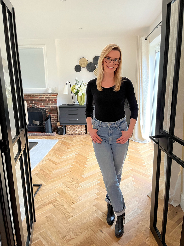 Smiling woman stood in modern living room with golden coloured herringbone flooring and brick feature fireplace.
